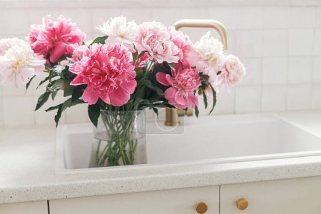Téléchargez les photos : Belles pivoines dans un vase en évier sur fond de robinet en laiton et comptoir blanc dans une nouvelle maison scandinave. Fleurs pivoine rose dans la cuisine moderne intérieur, arrangement floral d'été - en image libre de droit