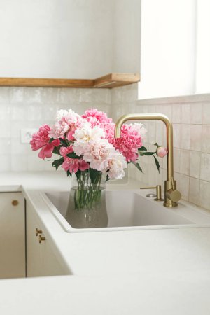 Photo for Beautiful peonies in vase in sink on background of brass faucet and window in new scandinavian house. Pink peony flowers in modern kitchen interior, summer floral arrangement - Royalty Free Image