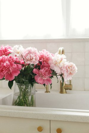 Photo for Beautiful peonies in vase in sink on background of brass faucet and window in new scandinavian house. Pink peony flowers in modern kitchen interior, summer floral arrangement - Royalty Free Image