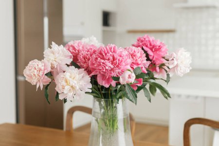 Téléchargez les photos : Belles pivoines dans un vase sur une table en bois sur fond de cuisine blanche élégante avec appareils électroménagers dans une nouvelle maison scandinave. Cuisine moderne aménagement intérieur et floral d'été - en image libre de droit