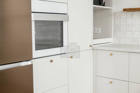 Téléchargez les photos : Design de cuisine minimaliste moderne. Élégantes armoires de cuisine blanches avec boutons en laiton, étagères en bois et appareils électroménagers dans une nouvelle maison scandinave. Intérieur de cuisine moderne - en image libre de droit