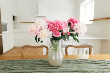 Téléchargez les photos : Belles pivoines dans un vase sur une table en bois sur fond de cuisine blanche élégante avec appareils électroménagers dans une nouvelle maison scandinave. Cuisine moderne aménagement intérieur et floral d'été - en image libre de droit