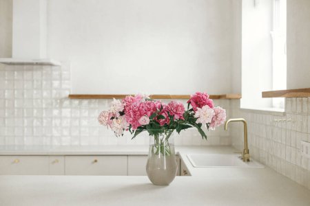 Téléchargez les photos : Belles pivoines dans un vase sur un comptoir en granit sur fond de cuisine blanche élégante avec des détails en laiton dans une nouvelle maison scandinave. Cuisine moderne aménagement intérieur et floral d'été - en image libre de droit