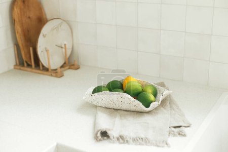 Photo for Limes in bowl and towel on background of modern white kitchen with granite countertop and white tiles in new scandinavian house. Modern kitchen interior - Royalty Free Image