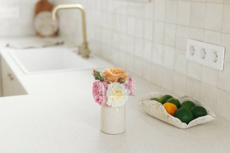 Téléchargez les photos : Belles roses et citrons verts dans un bol sur fond de cuisine blanche moderne avec robinet en laiton, comptoir en granit, étagères en bois avec ustensiles dans une nouvelle maison scandinave. Intérieur de cuisine moderne - en image libre de droit