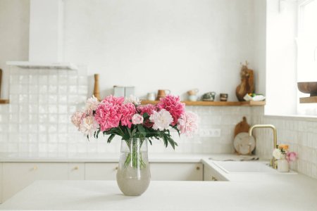 Téléchargez les photos : Belles pivoines dans un vase sur comptoir en granit sur fond de cuisine blanche élégante avec des étagères en bois et des appareils électroménagers dans une maison neuve. Intérieur de cuisine minimaliste moderne - en image libre de droit