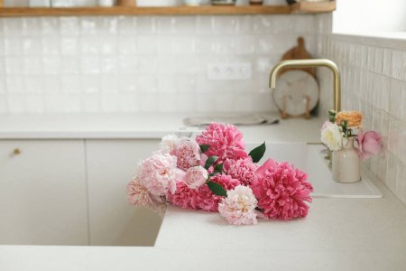 Téléchargez les photos : Pivoine rose et roses fleurs dans l'intérieur de la cuisine moderne, arrangement floral d'été dans la ferme. Belles pivoines dans l'évier sur fond de robinet en laiton et comptoir blanc dans une nouvelle maison scandinave - en image libre de droit