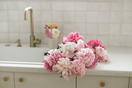 Téléchargez les photos : Belles pivoines dans l'évier sur fond de robinet en laiton et comptoir blanc dans une nouvelle maison scandinave. Pivoine rose et roses fleurs dans l'intérieur de la cuisine moderne, arrangement floral d'été - en image libre de droit