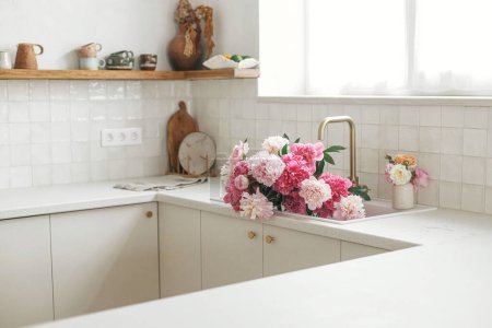 Photo for Pink peony and roses flowers in modern kitchen interior, summer floral arrangement. Beautiful peonies in sink on background of brass faucet and white counter in new scandinavian house - Royalty Free Image
