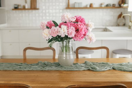Photo for Beautiful peonies in vase on wooden table on background of stylish white kitchen with appliances in new rustic farmhouse. Modern kitchen interior. Minimal kitchen design - Royalty Free Image
