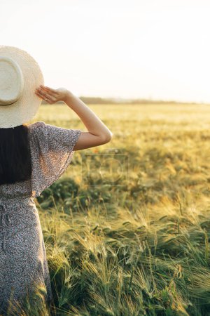 Photo for Beautiful woman in floral dress standing in barley field in sunset light. Stylish female holding straw hat and relaxing in evening summer countryside. Atmospheric moment, rustic slow life - Royalty Free Image