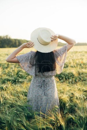 Photo for Beautiful woman in floral dress standing in barley field in sunset light. Stylish female holding straw hat and relaxing in evening summer countryside. Atmospheric moment, rustic slow life - Royalty Free Image