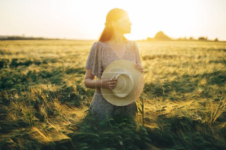 Photo for Beautiful woman in floral dress standing in barley field in sunset light. Atmospheric tranquil moment, rustic slow life. Stylish female holding straw hat and enjoying evening summer countryside - Royalty Free Image