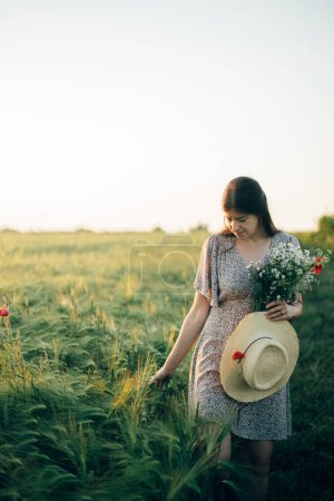 Foto de Hermosa mujer con flores silvestres y sombrero de paja caminando en el campo de cebada en la luz del atardecer. Elegante mujer relajándose en el campo de verano por la noche y recogiendo flores. Momento de tranquilidad atmosférica - Imagen libre de derechos