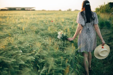 Téléchargez les photos : Belle femme avec fleurs sauvages et chapeau de paille marchant dans le champ d'orge à la lumière du coucher du soleil. Femme élégante relaxant en soirée campagne d'été et cueillette de fleurs. Moment de tranquillité atmosphérique - en image libre de droit