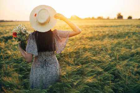 Téléchargez les photos : Belle femme au chapeau avec bouquet de fleurs sauvages debout dans le champ d'orge à la lumière du coucher du soleil. Femme élégante relaxant en soirée campagne d'été et cueillette de fleurs. Moment de tranquillité atmosphérique - en image libre de droit
