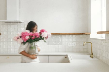 Téléchargez les photos : Femme heureuse floue arrangeant des pivoines dans un vase sur fond de cuisine blanche minimale dans une nouvelle maison moderne. Image de mouvement de la maison de décoration de femme au foyer avec des fleurs, entretien ménager - en image libre de droit