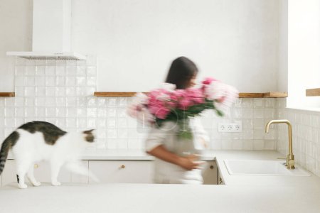 Photo for Blurred woman with her cat arranging peonies in vase on background of minimal white kitchen in new modern home. Motion image of housewife decorating house with flowers, housekeeping - Royalty Free Image
