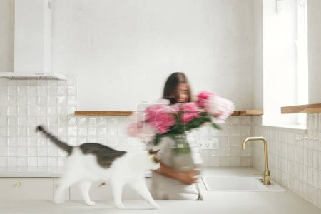 Foto de Mujer borrosa con su gato arreglando peonías en jarrón en el fondo de la cocina blanca mínima en el nuevo hogar moderno. Imagen en movimiento de ama de casa decorando la casa con flores, limpieza - Imagen libre de derechos