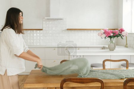 Foto de Mujer arreglando corredor de mesa textil verde en mesa de madera en el nuevo hogar moderno. Joven mujer decorando el comedor en el fondo de la cocina blanca mínima, mudanza y limpieza - Imagen libre de derechos