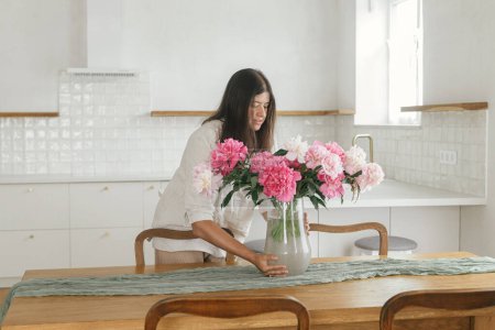 Foto de Mujer arreglando hermosas peonías en jarrón en un nuevo hogar moderno. Joven casa de decoración femenina con flores en el fondo de la cocina mínima blanca, moviéndose en casa escandinava - Imagen libre de derechos