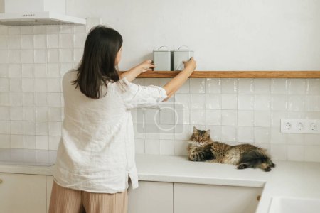 Photo for Stylish woman with her cat unpacking and decorating shelves with utensils in new minimal white kitchen. Housewife cleaning up kitchen after moving in with cute pet in new modern home - Royalty Free Image