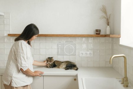 Foto de Mujer con estilo acariciando a su gato en la nueva cocina mínima blanca. Ama de casa limpiar la cocina después de mudarse con una linda mascota en un nuevo hogar moderno - Imagen libre de derechos