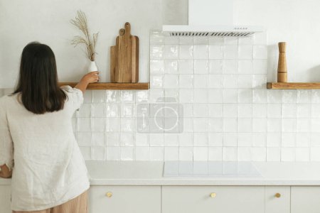 Téléchargez les photos : Femme élégante déballer et décorer des étagères avec des ustensiles dans la nouvelle cuisine blanche minimale. Femme au foyer nettoyer la cuisine après avoir déménagé dans une nouvelle maison scandinave moderne - en image libre de droit