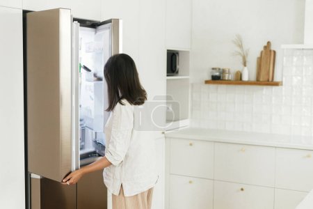 Téléchargez les photos : Femme élégante ouvrant réfrigérateur et regardant à l'intérieur dans la nouvelle cuisine blanche minimale. Femme au foyer nettoyer la cuisine dans une nouvelle maison scandinave moderne. Concept alimentaire et diététique - en image libre de droit