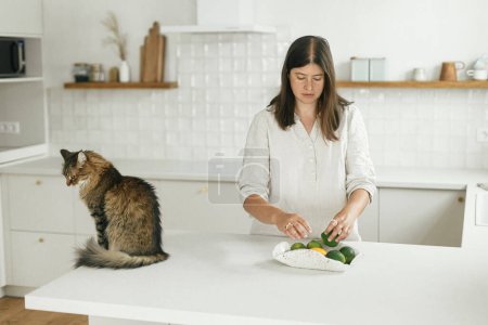 Foto de Mujer con estilo y su gato en la nueva cocina mínima blanca. Ama de casa limpiar la cocina después de mudarse con una linda mascota en un nuevo hogar moderno - Imagen libre de derechos