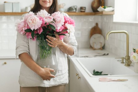 Photo for Woman holding beautiful peonies in vase at sink with brass faucet and granite countertop. Stylish female holding pink peony flowers in new modern kitchen, housewife decorating new home - Royalty Free Image