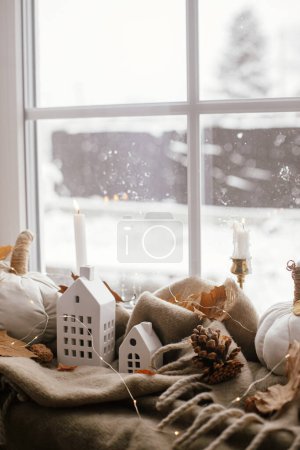 Foto de Autumn still life. Cozy pumpkin pillows, fall leaves, candle, lights and cute buildings decoration on brown scarf on windowsill. Fall home decor. Happy Thanksgiving - Imagen libre de derechos