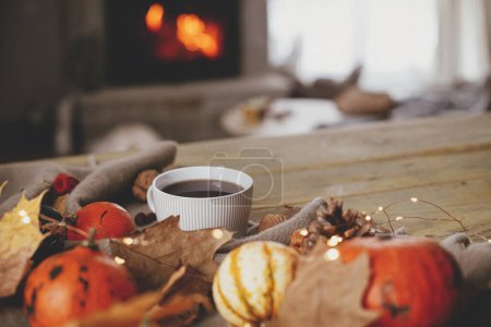Photo for Warm cup of tea, pumpkins, autumn leaves, cones, cozy scarf and lights on rustic wooden table in farmhouse. Fall in rural home. Happy Thanksgiving. Fall hygge still life, copy space - Royalty Free Image