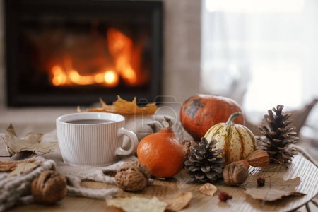 Téléchargez les photos : Cozy autumn. Warm cup of tea, pumpkins, autumn leaves, cones, cozy scarf on rustic wooden table on background of fireplace in farmhouse. Comfort fall in rural home. Happy Thanksgiving - en image libre de droit