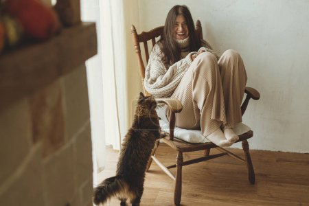 Foto de Mujer feliz en suéter de punto acogedor jugando con el gato mientras está sentado en la silla de madera vintage. Acogedor otoño con mascota en casa. Elegante hembra y adorable gato tabby en granja - Imagen libre de derechos