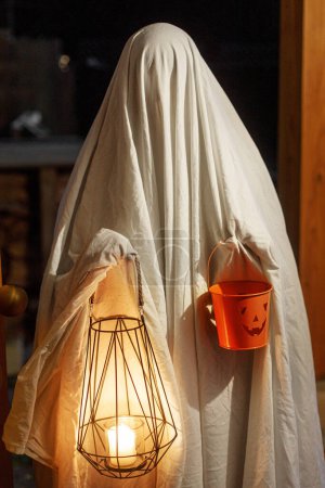 Photo for Trick or treat! Spooky ghost holding glowing lantern and halloween candy bucket in doors in dark. Scary atmospheric halloween time, person dressed as ghost trick or treating. Happy Halloween - Royalty Free Image