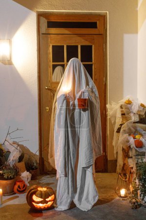 Téléchargez les photos : Fantôme effrayant avec un seau de bonbons d'Halloween et une lanterne Jack o aux portes dans une nuit effrayante. Joyeux Halloween ! Personne habillée comme tour de fantôme ou traiter sur le porche avec des décorations d'Halloween - en image libre de droit