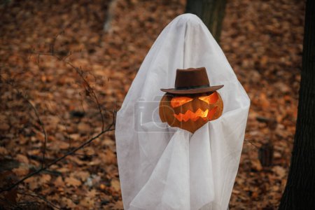 Téléchargez les photos : Élégant fantôme tenant Jack o lanterne dans la forêt d'automne humide. Personne habillée en drap blanc comme fantôme avec citrouille debout dans les bois d'automne du soir. Joyeux Halloween ! Trick ou traiter - en image libre de droit