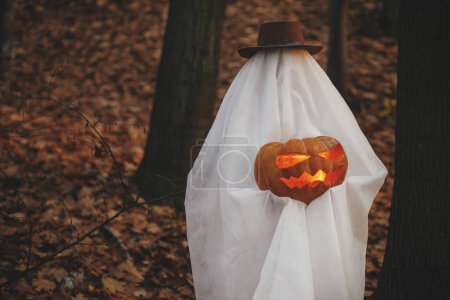 Foto de ¡Feliz Halloween! Fantasma elegante sosteniendo jack o linterna en el bosque de otoño malhumorado. Persona vestida con sábana blanca como fantasma con calabaza de pie en los bosques de otoño por la noche. Truco o trato - Imagen libre de derechos