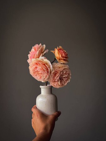 Foto de Mano sosteniendo hermosas rosas pastel en jarrón sobre fondo rústico. Flores con estilo naturaleza muerta, composición artística. Papel pintado vertical floral - Imagen libre de derechos