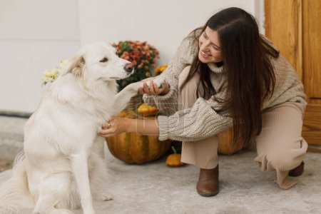 Photo for Happy woman in knitted sweater playing with cute dog at house entrance decorated with autumn pumpkins, pots with chrysanthemums and heather. Owner with pet and fall arrangement - Royalty Free Image