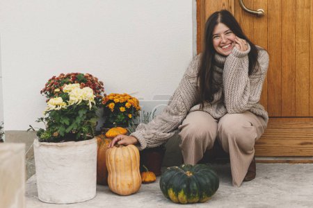 Téléchargez les photos : Femme heureuse en pull tricoté décorant l'entrée de la maison avec des citrouilles d'automne, des pots avec des chrysanthèmes et de la bruyère. Décoration automnale élégante de la porte d'entrée de la ferme. Arrangement d'automne - en image libre de droit