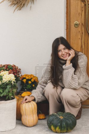 Téléchargez les photos : Femme heureuse en pull tricoté décorant l'entrée de la maison avec des citrouilles d'automne, des pots avec des chrysanthèmes et de la bruyère. Décoration automnale élégante de la porte d'entrée de la ferme. Arrangement d'automne - en image libre de droit