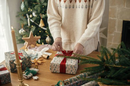 Téléchargez les photos : Femme enveloppant cadeau de Noël élégant avec ruban rouge sur une table en bois avec des décorations festives dans une chambre scandinave décorée. Joyeux Noël et Joyeuses Fêtes ! Tenue de main présente - en image libre de droit