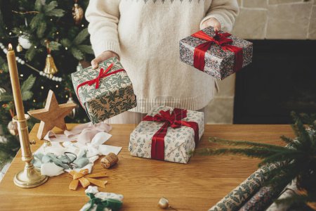 Foto de Mujer sosteniendo elegantes regalos de Navidad con cinta roja en la mesa de madera con decoraciones festivas en el fondo del árbol decorado y la chimenea en la habitación escandinava. Feliz Navidad.! - Imagen libre de derechos