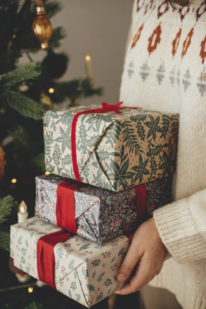 Foto de Mujer en suéter con elegantes regalos de Navidad con cinta roja en el moderno árbol vintage decorado en la habitación escandinava. Feliz Navidad y Felices Fiestas! - Imagen libre de derechos