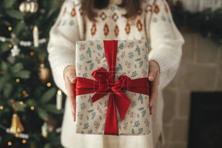 Foto de Feliz Navidad y Felices Fiestas! Elegante regalo de Navidad con cinta roja en las manos de la mujer de cerca en el moderno árbol vintage decorado en la habitación escandinava - Imagen libre de derechos