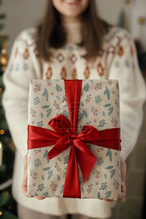 Foto de Elegante regalo de Navidad con cinta roja en las manos de la mujer de cerca en el moderno árbol vintage decorado en la habitación escandinava. Feliz Navidad y Felices Fiestas! - Imagen libre de derechos