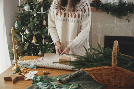 Téléchargez les photos : Femme en pull élégant enveloppant cadeau de Noël en papier sur une table en bois avec des décorations festives dans une chambre scandinave décorée. Joyeux Noël ! Emballage des mains présent gros plan - en image libre de droit