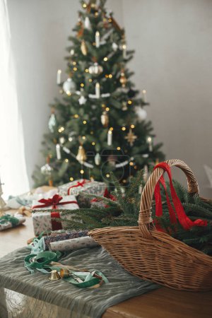 Photo for Rustic basket with fir branches, stylish christmas gifts and festive decorations on table on background of beautiful vintage christmas tree and fireplace. Merry Christmas and Happy Holidays! - Royalty Free Image